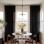 A dining room scene with tall black curtains hanging on a window, a round white table, four wooden chairs, a pendant light, and a potted tree in a bright interior.