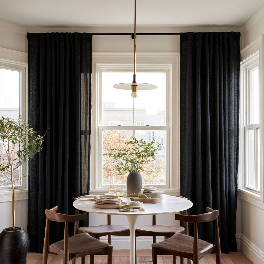 A dining room scene with tall black curtains hanging on a window, a round white table, four wooden chairs, a pendant light, and a potted tree in a bright interior.