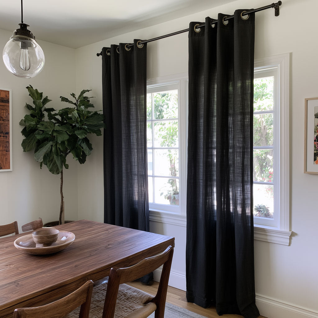 Black sheer grommet curtains hanging on a black rod over two white-framed windows in a dining room with a wooden table and chairs.
