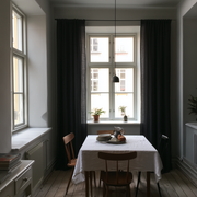A dining nook with a white tablecloth on a wooden table, surrounded by wooden chairs, set against a window with dark charcoal curtains and a hanging pendant light.