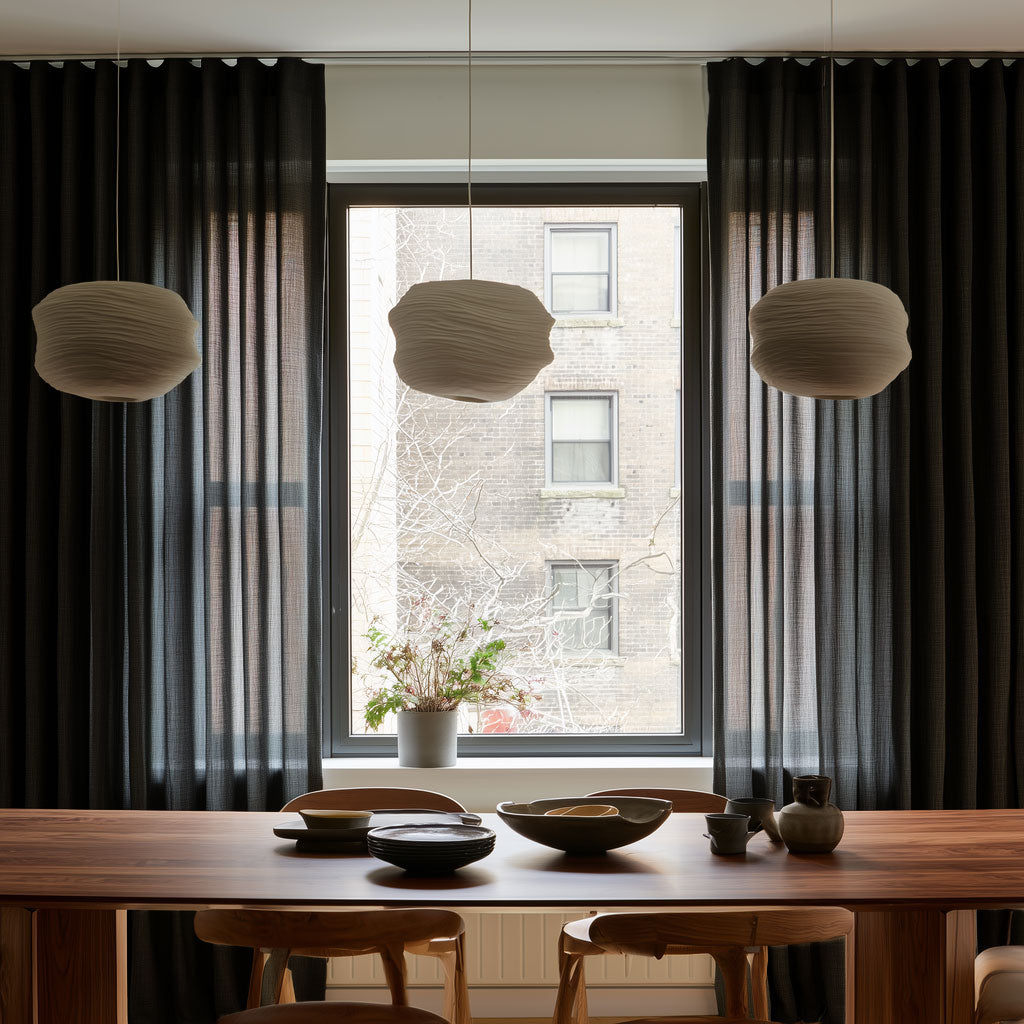 A styled dining room interior with a wooden dining table, chairs, three textured pendant lights, dark charcoal curtains, and a large window looking out onto a brick building.