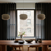 A styled dining room interior with a wooden dining table, chairs, three textured pendant lights, dark charcoal curtains, and a large window looking out onto a brick building.
