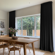 A pair of floor-length black pleated curtains hanging on either side of a large white-framed window in a modern dining room with a wooden table and chairs.