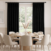 Black curtain panels hanging on a dark curtain rod in a bright dining room with a round wooden table and beige upholstered chairs.