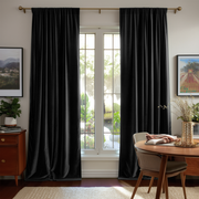 A pair of floor-length black curtain panels hanging on a gold rod over a tall white-framed window in a styled room with framed wall art, a wooden side table, and a dining table with a chair.