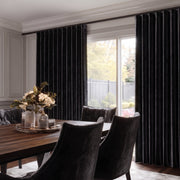 Black pleated window curtains hanging on a dark rod in a dining room setting with a wooden table and dark upholstered chairs.