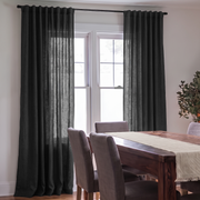 A pair of dark charcoal gray grommet-top curtains hanging on a black rod in front of a white-framed window, shown in a dining room setting with a wooden table and gray chairs.
