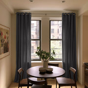 A pair of dark slate blue grommet-top window curtains hanging on a silver rod in a dining room setting with a wooden table and chairs.