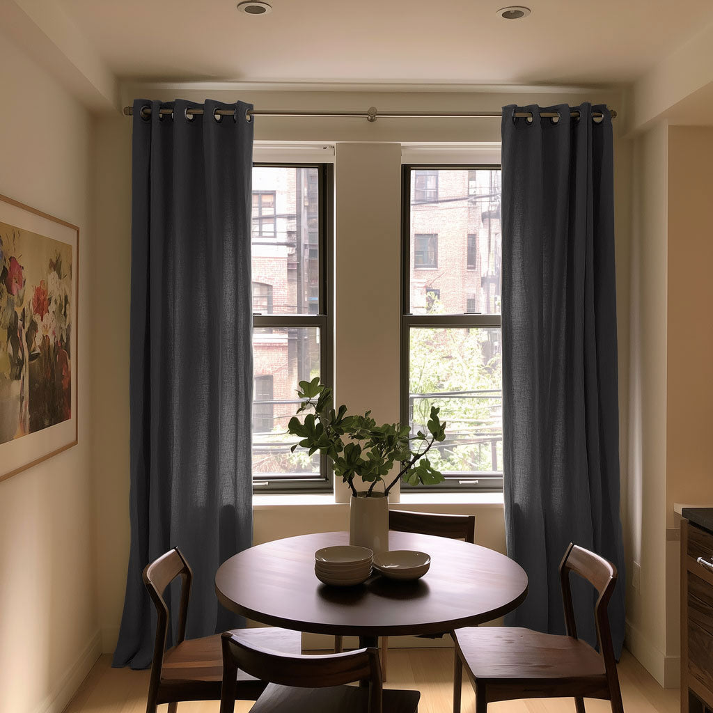 A pair of dark slate blue grommet-top window curtains hanging on a silver rod in a dining room setting with a wooden table and chairs.