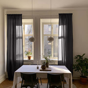 A dining room scene with tall sheer black curtains hanging on both sides of two tall windows, with pendant lights, a white tablecloth, chairs, and potted plants in a bright interior.