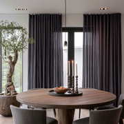 A pair of floor-length dark charcoal gray curtains hanging on a black-framed window in a modern dining room with a round wooden table and gray chairs.