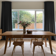A modern dining room setup featuring a light oak wood rectangular table and four matching mid-century style chairs with brown upholstered seats, set on a textured cream rug in front of a large window with dark gray curtains.