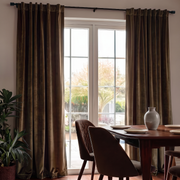 A pair of floor-length brown velvet curtains hanging on a black rod over a white French door, with a dining table and chairs in a sunlit room.