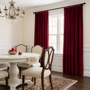 Deep burgundy velvet window curtains hanging on a black rod in a dining room setting with a white table, wooden chairs, and a patterned rug