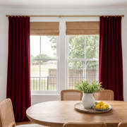 A pair of deep burgundy curtain panels hanging on a gold rod with matching woven jute roman blinds in front of a white-framed window, with a wooden dining table and chairs visible in the foreground.