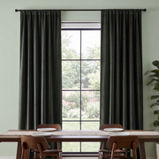 Dark charcoal gray curtain panels hanging on a black rod over a tall window in a dining room setting with a wooden table and chairs.