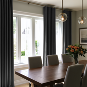A dining room scene with dark charcoal gray curtains hanging around tall white-framed windows, a wooden dining table, upholstered chairs, pendant lights, and a vase of flowers.