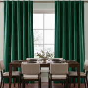 A pair of deep emerald green curtain panels hanging on a rod over a window, flanking a dining table with beige upholstered chairs in a bright, modern dining room.