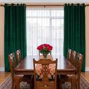 A pair of deep emerald green velvet grommet curtains hanging on a wooden rod in a dining room setting with a wooden dining table, chairs, and a vase of red roses.