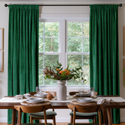 A pair of deep emerald green curtain panels hanging on a black rod over a tall white window, styled in a dining room with a wooden table, chairs, and a floral centerpiece.