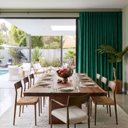 A long wooden dining table set with white plates, silver cutlery, and a floral centerpiece, surrounded by matching wooden chairs with woven seats. The dining area is styled with a large emerald green curtain, a potted plant, and a light rug, with an open sliding door revealing a backyard pool and patio in the background.