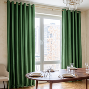 A pair of deep forest green grommet curtains hanging on a gold rod in a dining room setting with a wooden table, chairs, and a window view.