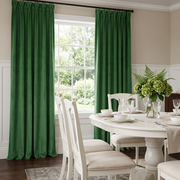A pair of deep forest green curtain panels hanging on a dark rod in a bright dining room with a white table, chairs, and a window behind them.