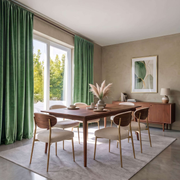 A dining room scene with a wooden dining table and six upholstered chairs arranged on a light rug, set in a modern room with green curtains, beige walls, and a sideboard.