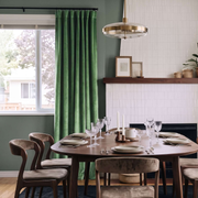 A styled dining room scene featuring a pair of forest green velvet curtains hanging next to a window with a view of trees outside. The room has a round wooden dining table set with plates and wine glasses, matching wooden chairs, a brass pendant light, and a white shiplap wall with a fireplace.