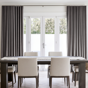 A pair of floor-length taupe gray curtains hanging on a rod over white French doors in a bright, modern dining room with a dark wood table and cream upholstered chairs.
