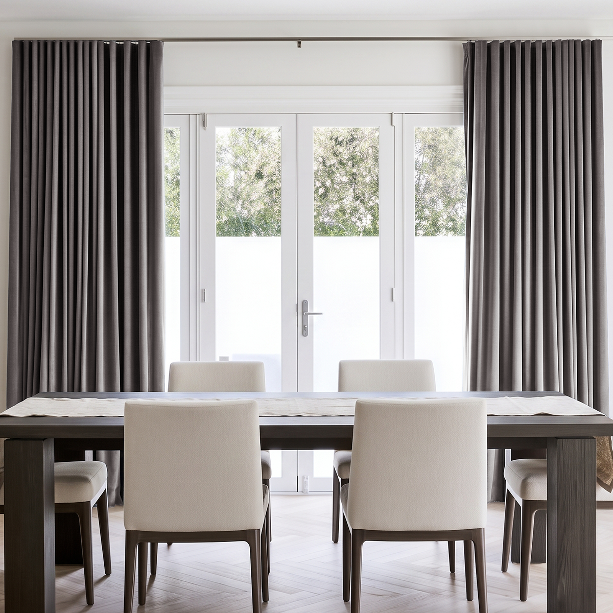 A pair of floor-length taupe gray curtains hanging on a rod over white French doors in a bright, modern dining room with a dark wood table and cream upholstered chairs.