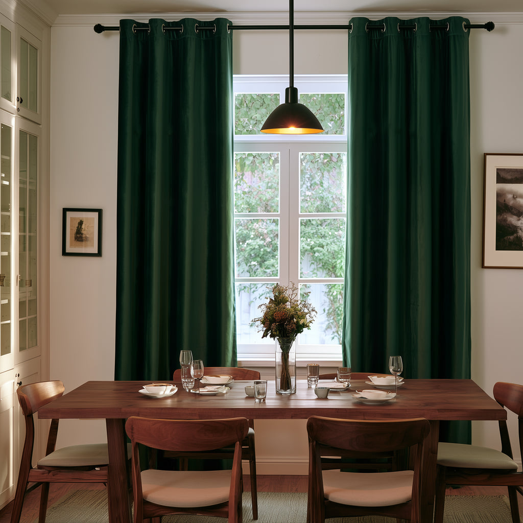 A pair of dark forest green grommet curtains hanging on a black rod in a dining room setting with a wooden table, chairs, and a window in the background.
