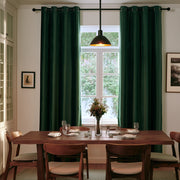 A pair of dark forest green grommet curtains hanging on a black rod in a dining room setting with a wooden table, chairs, and a window in the background.