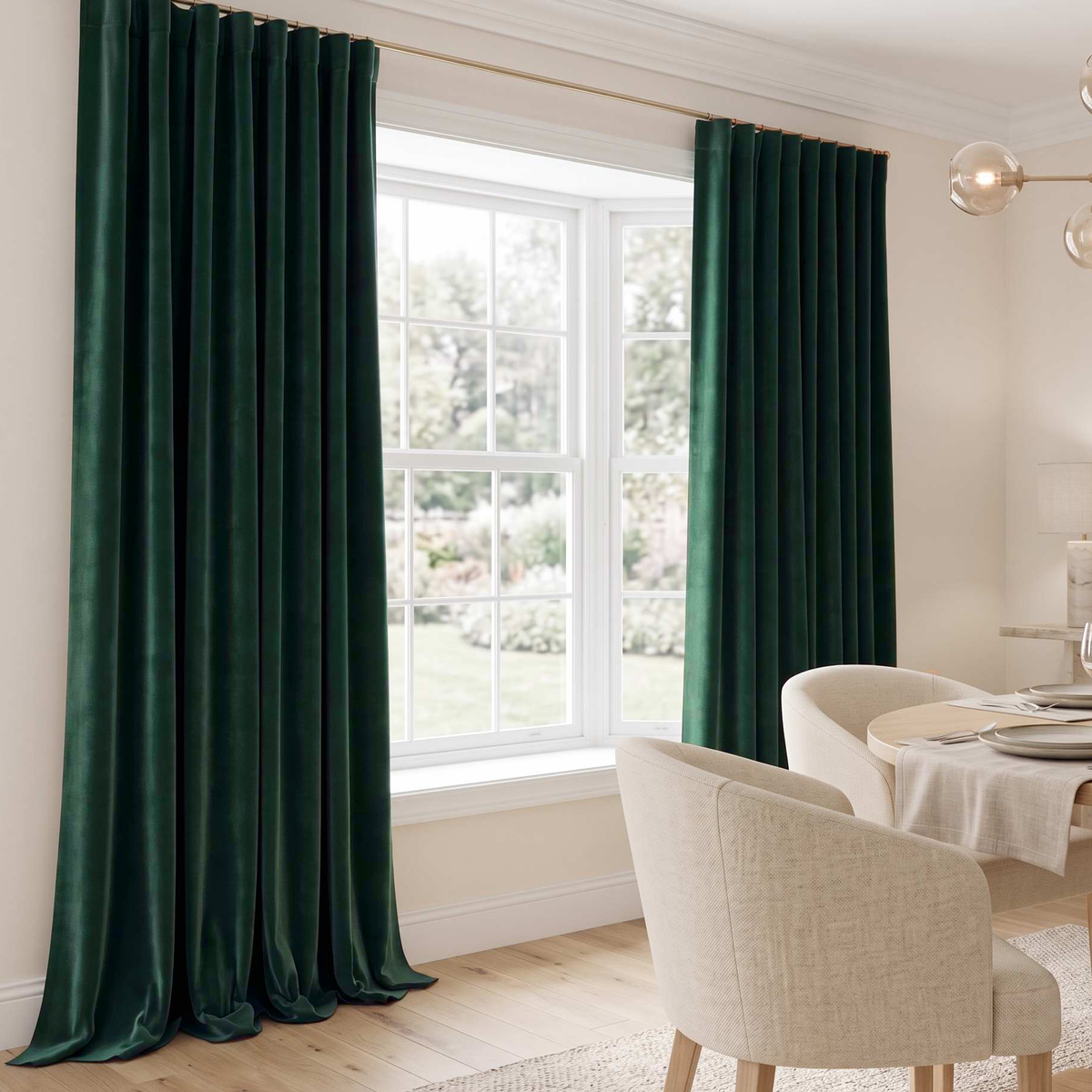 A pair of dark forest green velvet curtains hanging on a gold rod in a bright, modern room with a white window, light wood floors, and neutral-toned dining chairs.