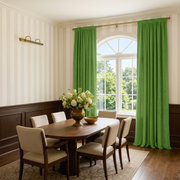 A dining room with a wooden table and six upholstered chairs, featuring a large arched window dressed in bright green curtains. The room has cream and beige vertical striped wallpaper, dark wood wainscoting, and a vase of white flowers on the table.