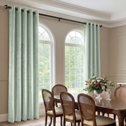 Light sage green curtain panels hanging on a black rod in a dining room with arched windows, a wooden dining table, and woven-back chairs.