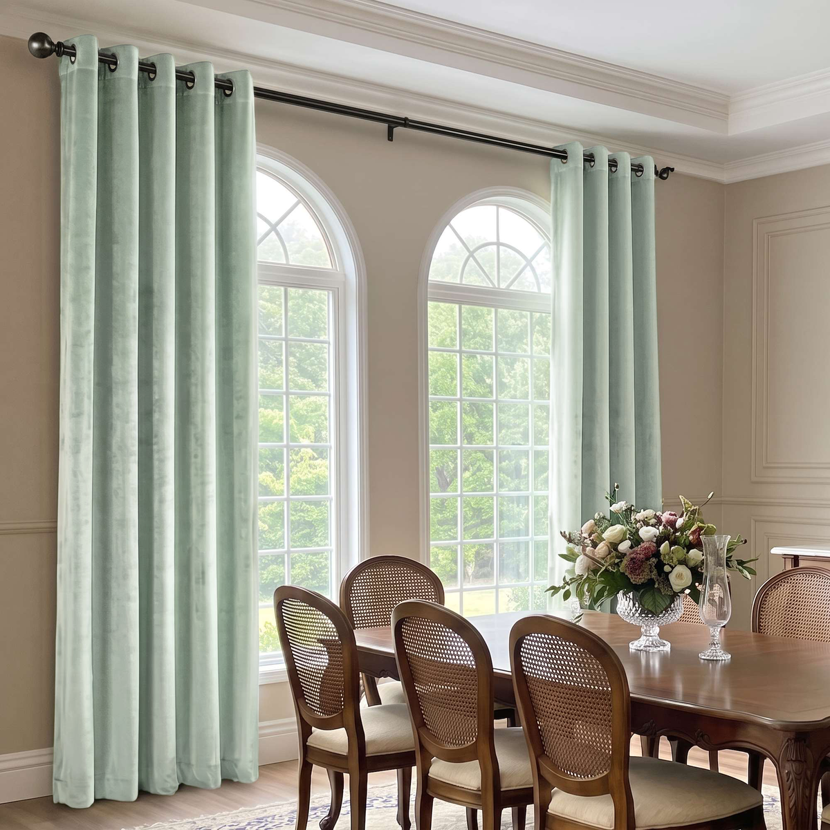 Light sage green curtain panels hanging on a black rod in a dining room with arched windows, a wooden dining table, and woven-back chairs.