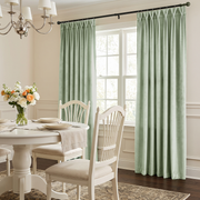 Light sage green curtain panels hanging on a black rod in a bright dining room with white chairs, a round table, and a window.