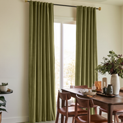 A pair of sage green curtain panels hanging on a black rod in a dining room setting with a wooden table and chairs.