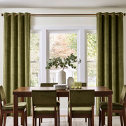 Olive green velvet grommet curtains hanging on a brass rod over a multi-pane window, flanking a wooden dining table and chairs in a bright, modern dining room.
