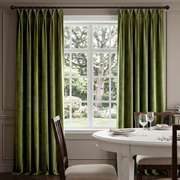 A pair of olive green velvet curtain panels hanging on a dark curtain rod in front of a white multi-pane window, with a dining table and chairs visible in the softly lit room.