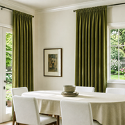 A pair of olive green pleated window curtains hanging on dark metal rods in a bright dining room with a white tablecloth and chairs.