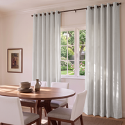 A pair of light sage green grommet curtains hanging on a dark metal rod in a dining room setting with a wooden table and white chairs.