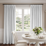 A pair of light gray pleated window curtains hanging on a black rod over a white multi-pane window, with a styled dining table and chairs in a bright room below.