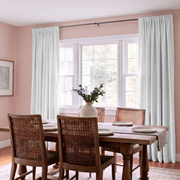 A dining room with a wooden table and woven chairs, featuring light sage green curtains hanging from a black rod over a multi-pane window. The walls are painted a soft dusty pink, and a white vase with greenery sits on the table.
