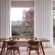 A pair of floor-length white pleated curtains hanging on a window in a dining room setting, with a wooden dining table and chairs visible in the foreground and a backyard garden outside.