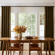 A dining room scene with a wooden table and chairs, a vase of autumn leaves on the table, and tall dark olive-brown curtains hanging in front of three large white-framed windows.