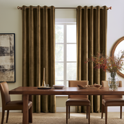 A pair of dark olive brown grommet-top curtains hanging on a brass rod over a window in a styled dining room with a wooden table and chairs.