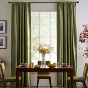 A pair of sage green curtain panels hanging on a black rod over a tall window in a dining room setting with a wooden table and chairs.