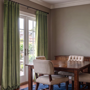 A pair of long sage green velvet curtains hanging on a black rod over a set of French doors in a dining room with a wooden table and upholstered chairs.